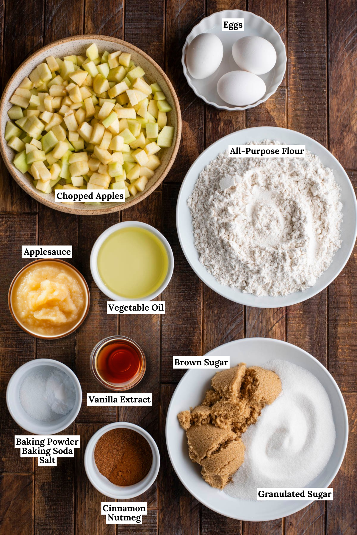 Ingredients for an Apple Bundt Cake are arranged on a wooden table: chopped apples, eggs, all-purpose flour, applesauce, vegetable oil, brown sugar, granulated sugar, vanilla extract, cinnamon, nutmeg, baking powder, baking soda, and salt.