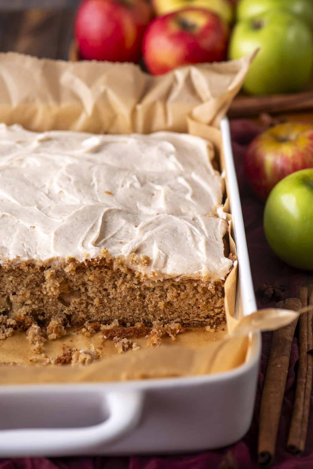 A white baking dish with a rectangular apple cake topped with creamy frosting, partially sliced. Whole red and green apples and cinnamon sticks are in the background. The cake sits on parchment paper.