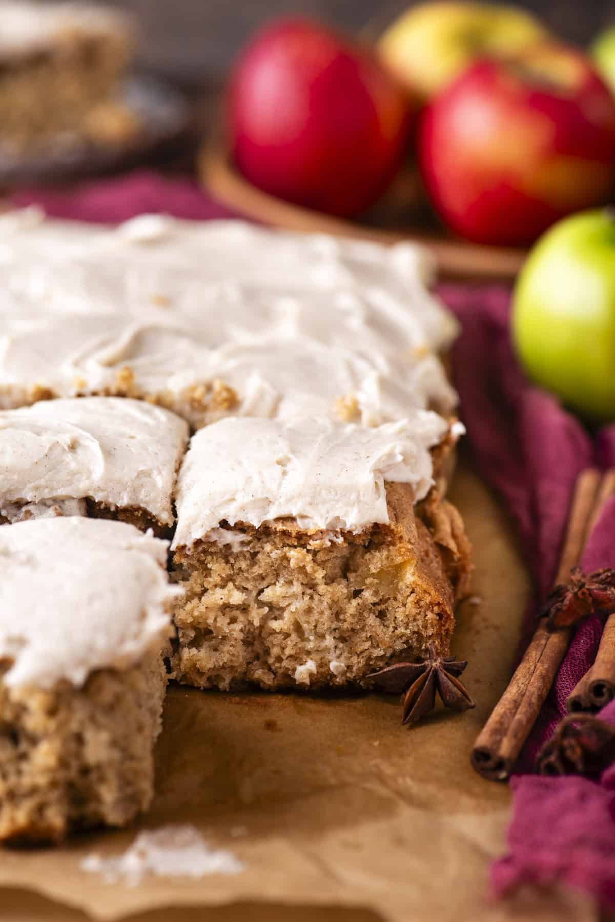 Frosted apple spice cake squares on parchment paper, with a bite taken from one piece. Cinnamon sticks, star anise, and whole apples are in the background, adding a cozy, autumn feel.