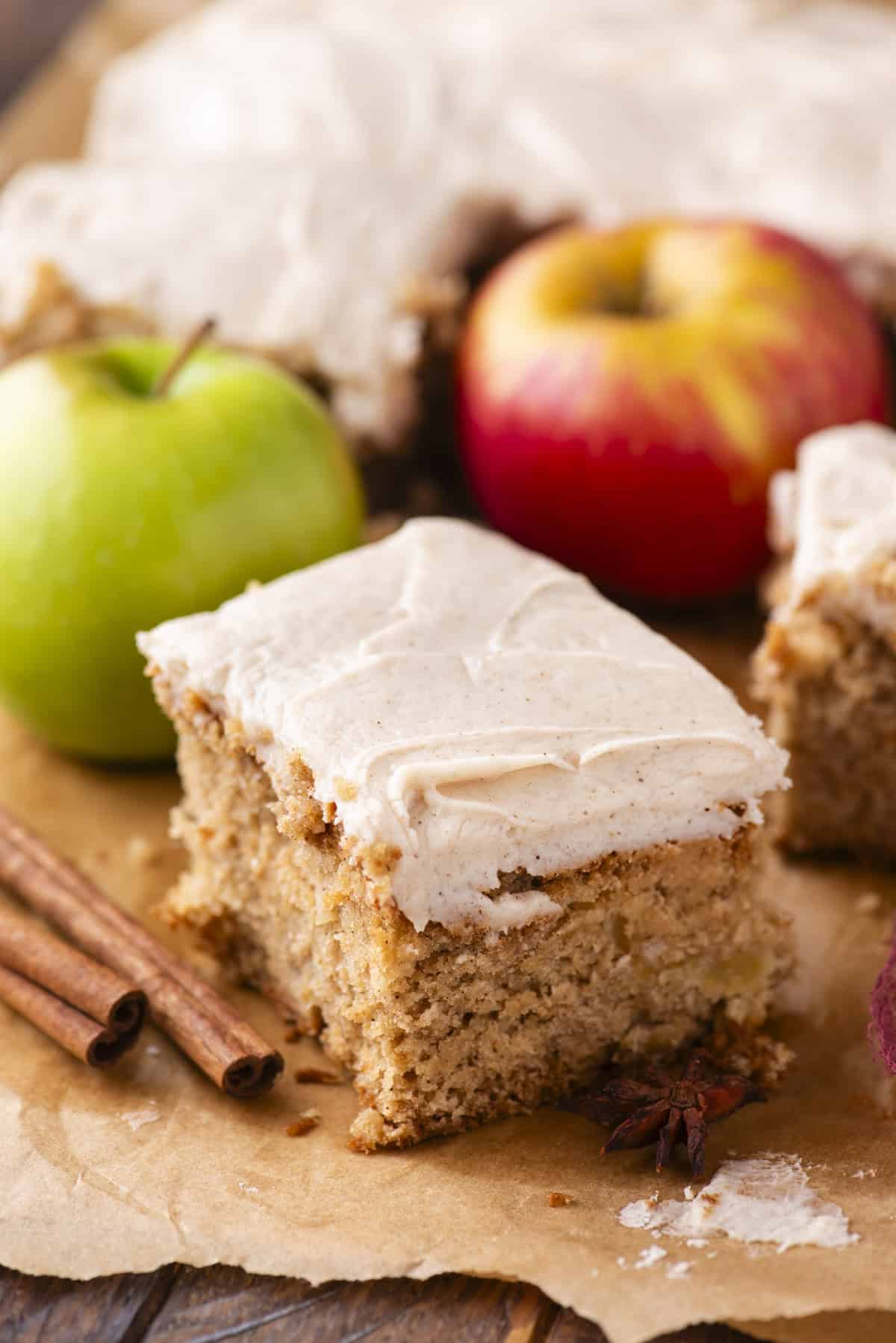 A slice of spiced apple cake with creamy frosting sits on parchment paper, surrounded by cinnamon sticks, a green apple, a red apple, and more cake in the background.