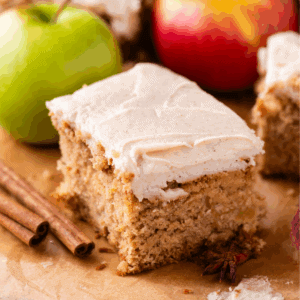 A slice of spiced apple cake topped with creamy frosting, surrounded by cinnamon sticks and whole apples on a parchment-lined surface.