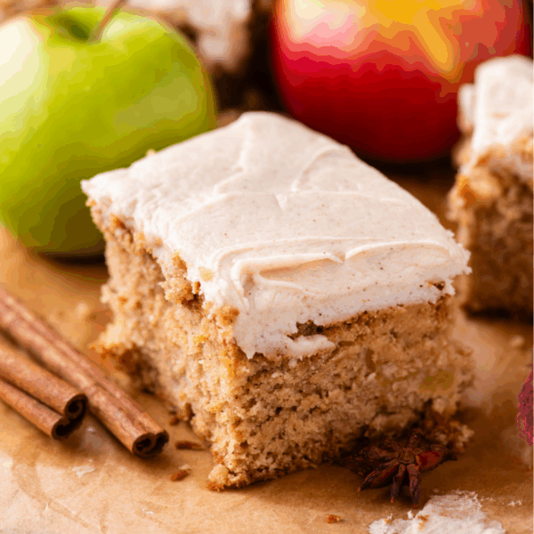 A slice of spiced apple cake topped with creamy frosting, surrounded by cinnamon sticks and whole apples on a parchment-lined surface.
