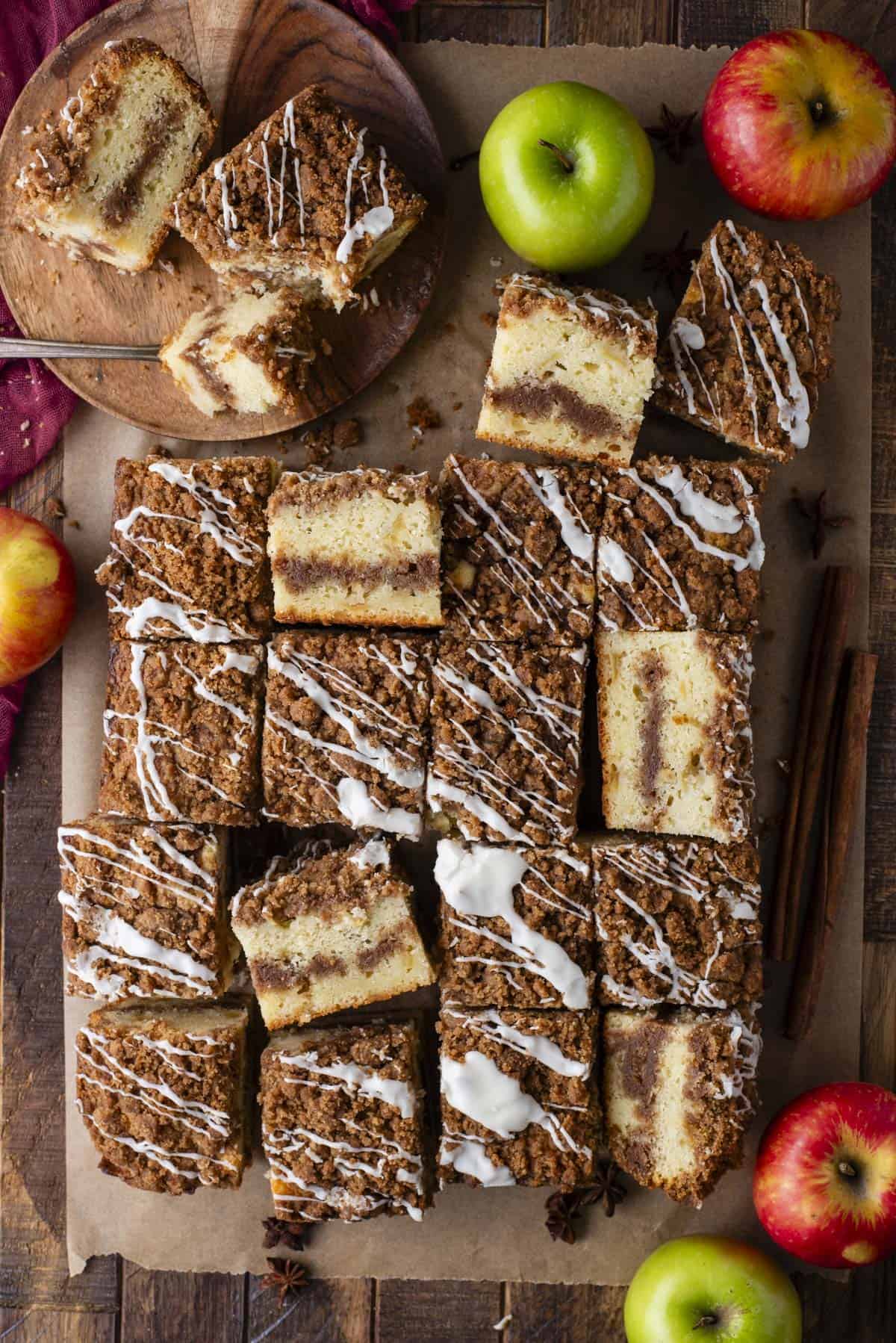 Overhead view of sliced apple coffee cake squares with crumb topping and white icing drizzle, arranged on parchment paper. Green and red apples, cinnamon sticks, and star anise are scattered around for decoration.