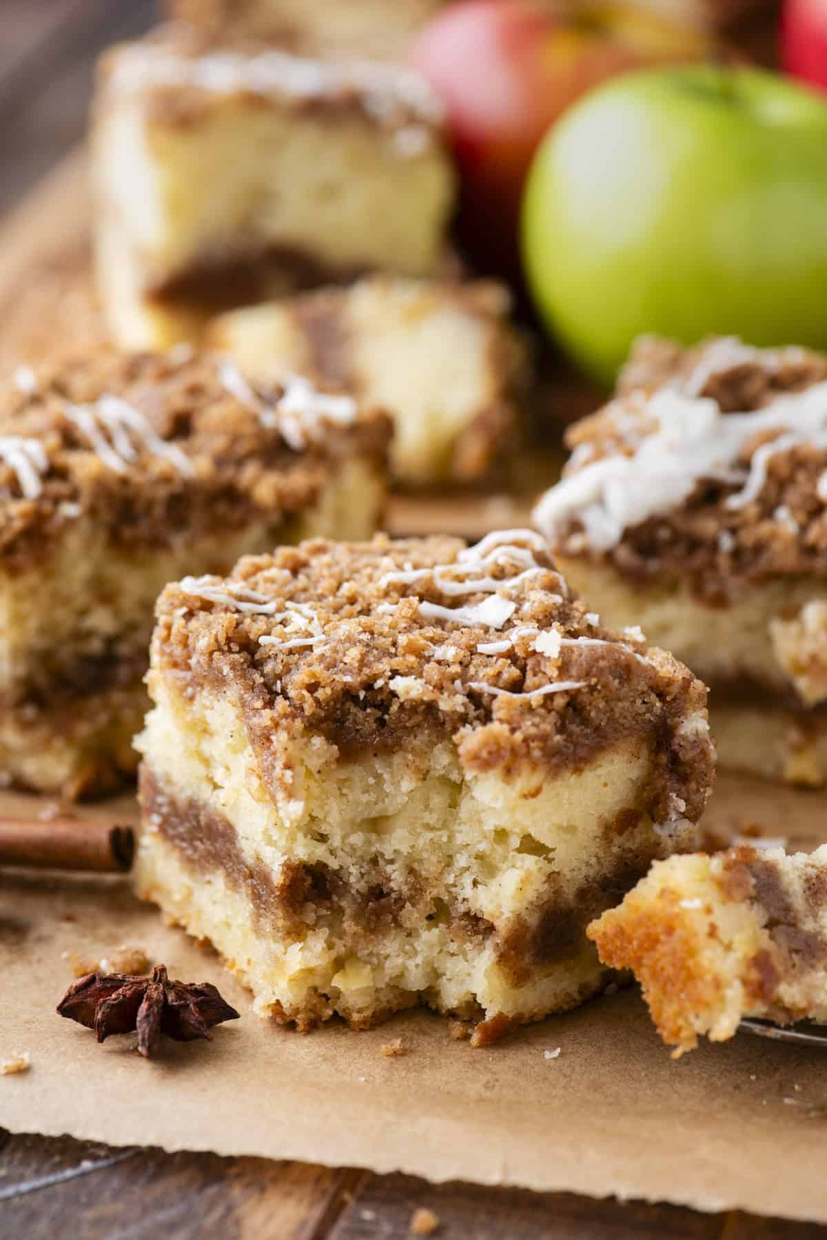 A close-up of several pieces of apple crumb cake on parchment paper, topped with a crumbly streusel and a light drizzle of icing, with fresh apples blurred in the background.