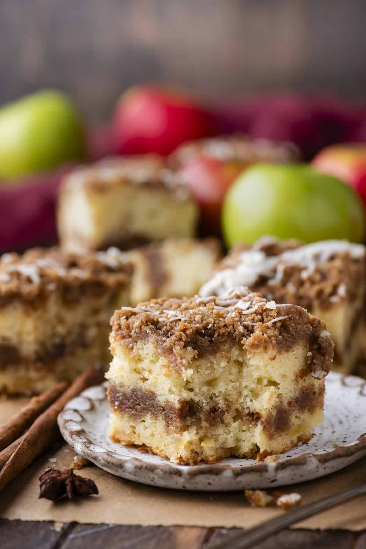A slice of crumbly coffee cake with cinnamon swirl on a plate, with more cake pieces and colorful apples blurred in the background.