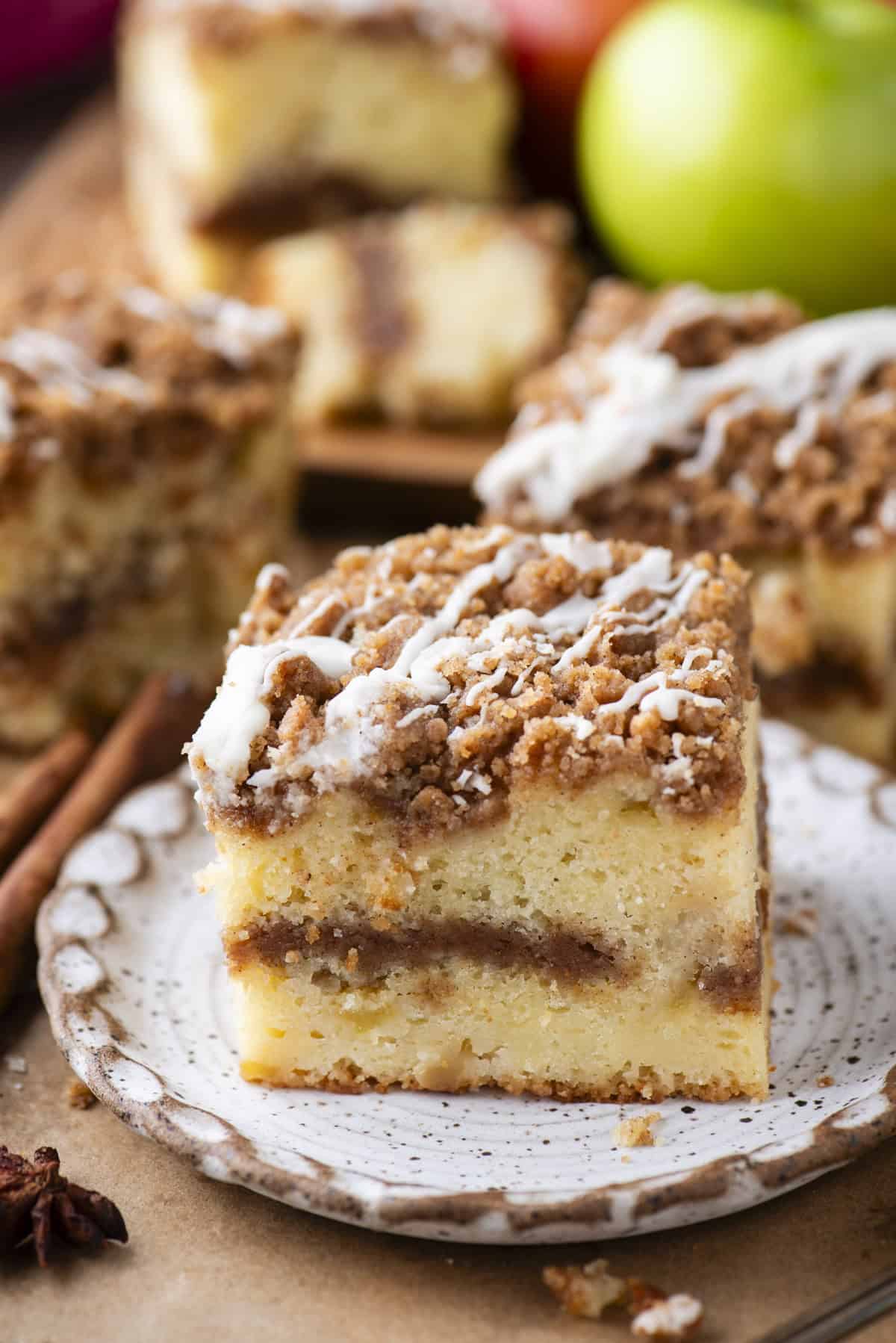 A slice of crumbly coffee cake with a cinnamon swirl and a drizzle of white icing sits on a decorative plate, with more cake pieces and green apples in the background.