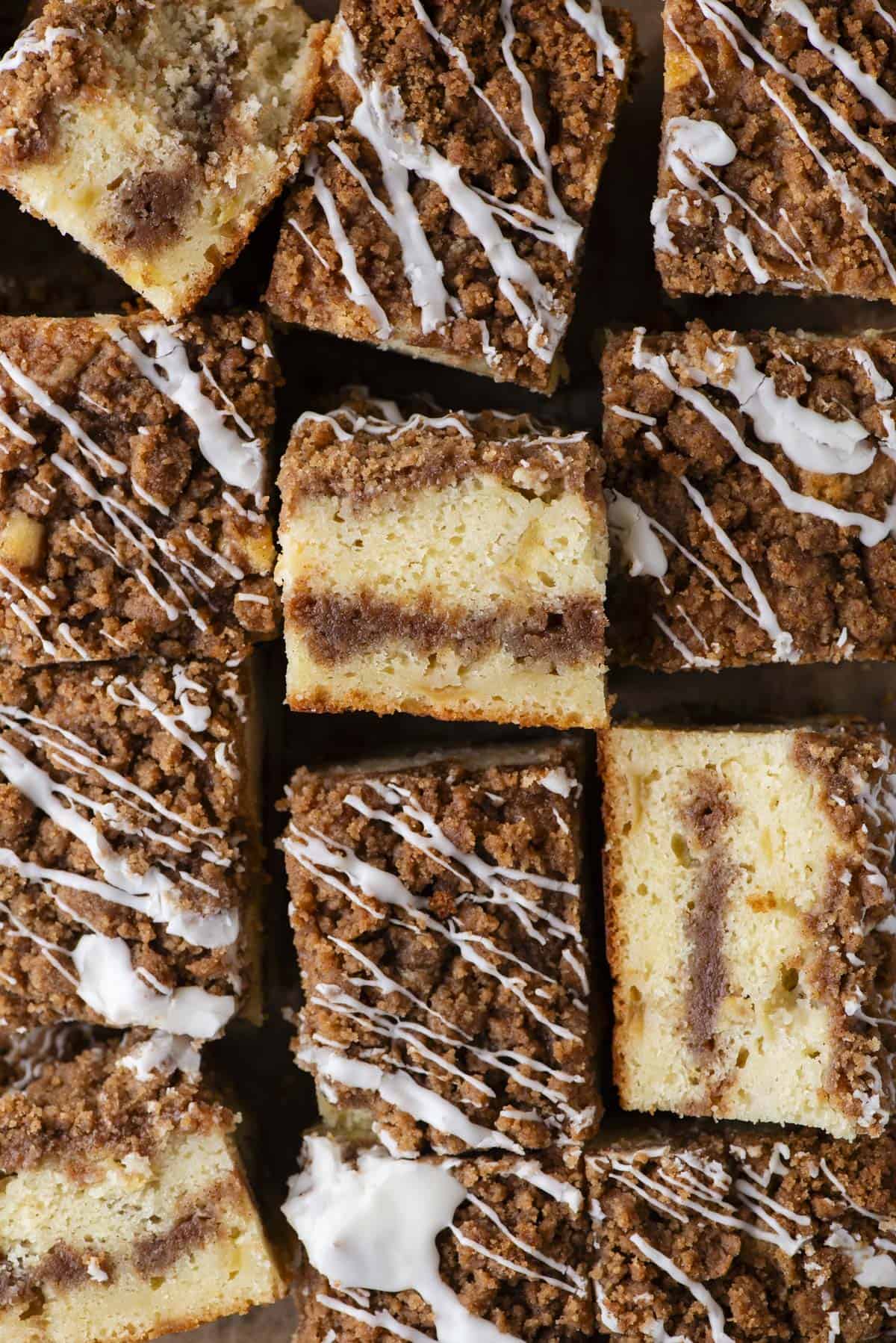 Close-up of several pieces of coffee cake with a crumbly cinnamon streusel topping and white icing drizzle, arranged closely together on a flat surface.