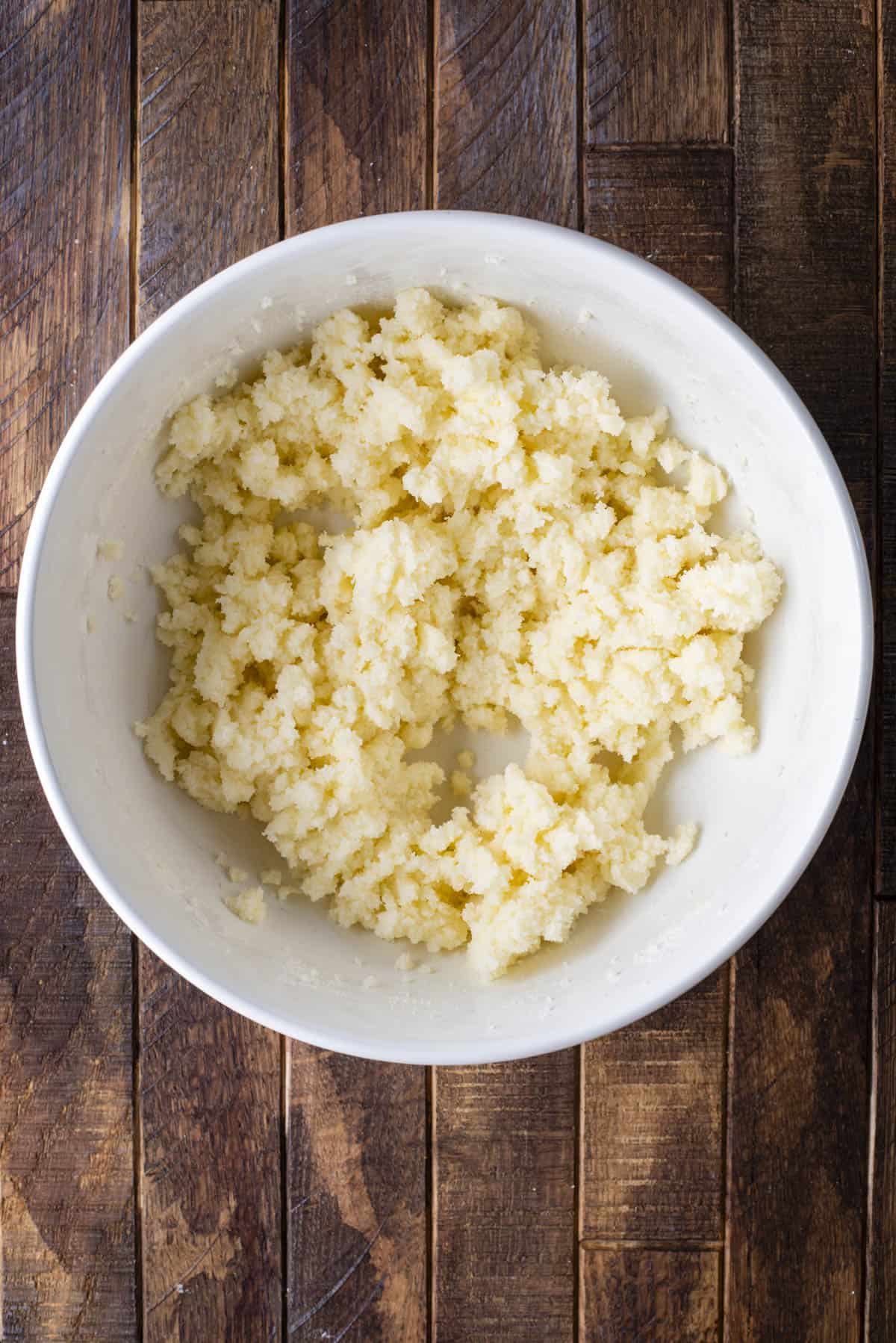 A white bowl containing a crumbly mixture of butter and sugar sits on a rustic wooden surface.
