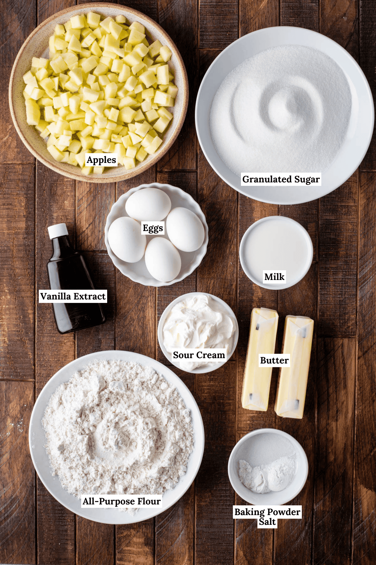 Top-down view of ingredients for baking: cubed apples, granulated sugar, eggs, milk, vanilla extract, sour cream, two sticks of butter, all-purpose flour, baking powder, and salt on a wooden surface, each labeled.