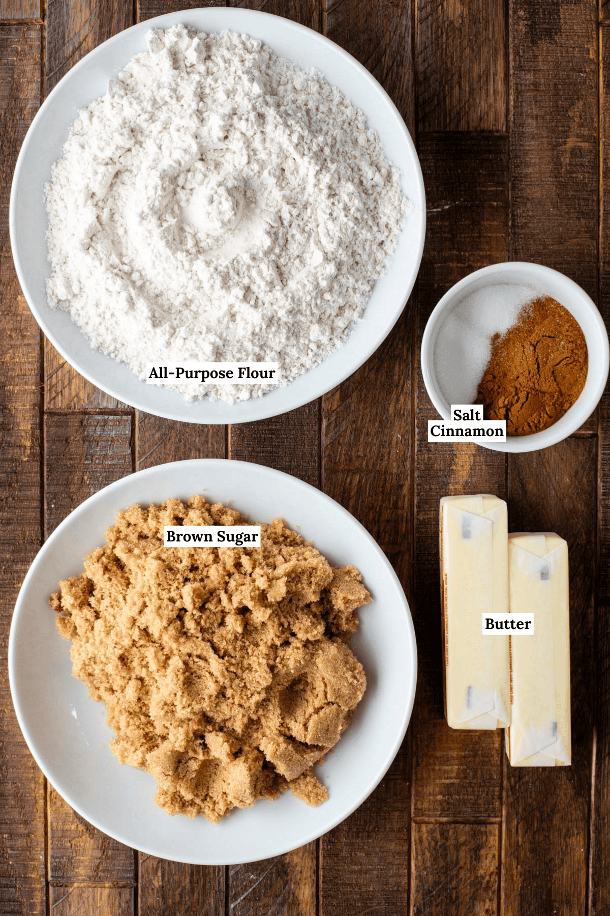 Overhead view of measured baking ingredients on a wooden surface: a bowl of all-purpose flour, a small bowl with salt and cinnamon, a plate of brown sugar, and two sticks of butter. Each item is labeled.