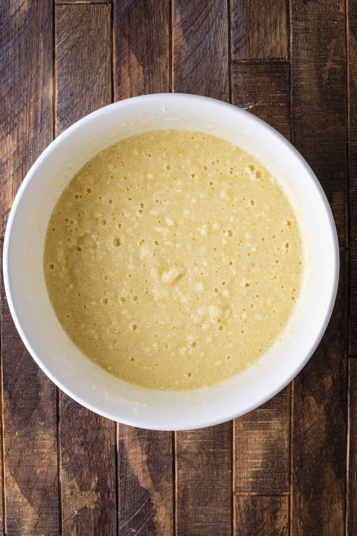 A white bowl filled with yellow pancake batter sits on a rustic wooden surface. The batter appears slightly lumpy and bubbly, ready to be cooked.