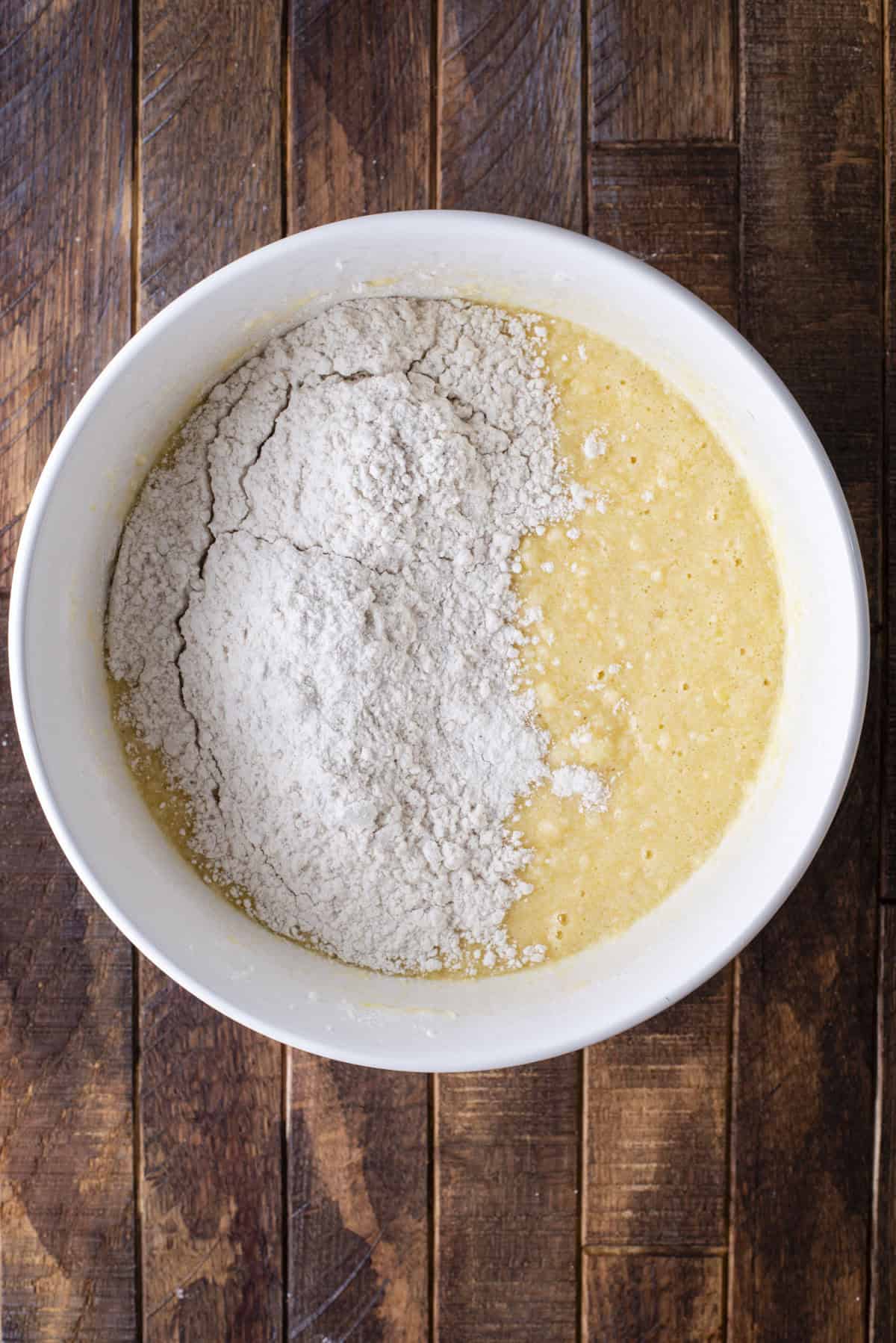 A white bowl contains yellow batter with a mound of white flour on top, sitting on a dark wooden surface.