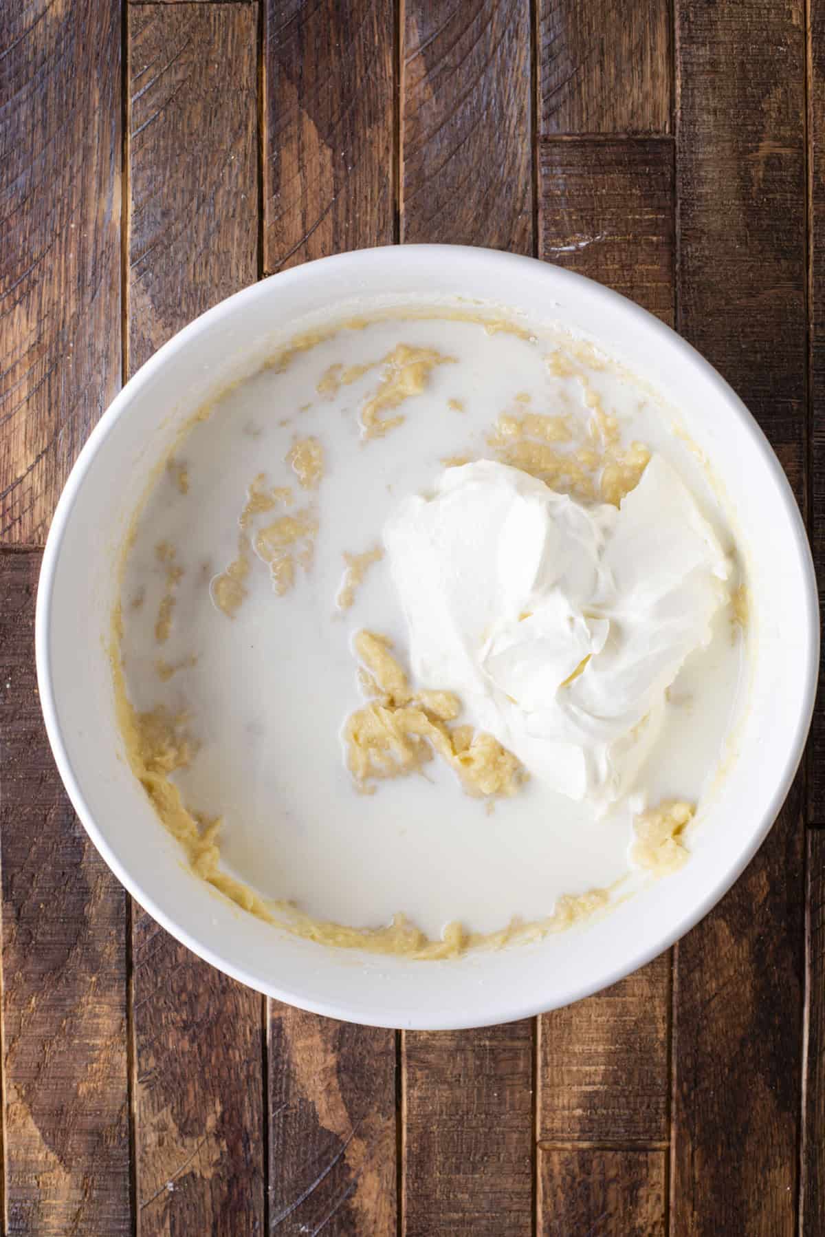 A white bowl on a wooden surface contains a mixture of milk, mashed bananas, and a large dollop of sour cream, partially blended together.