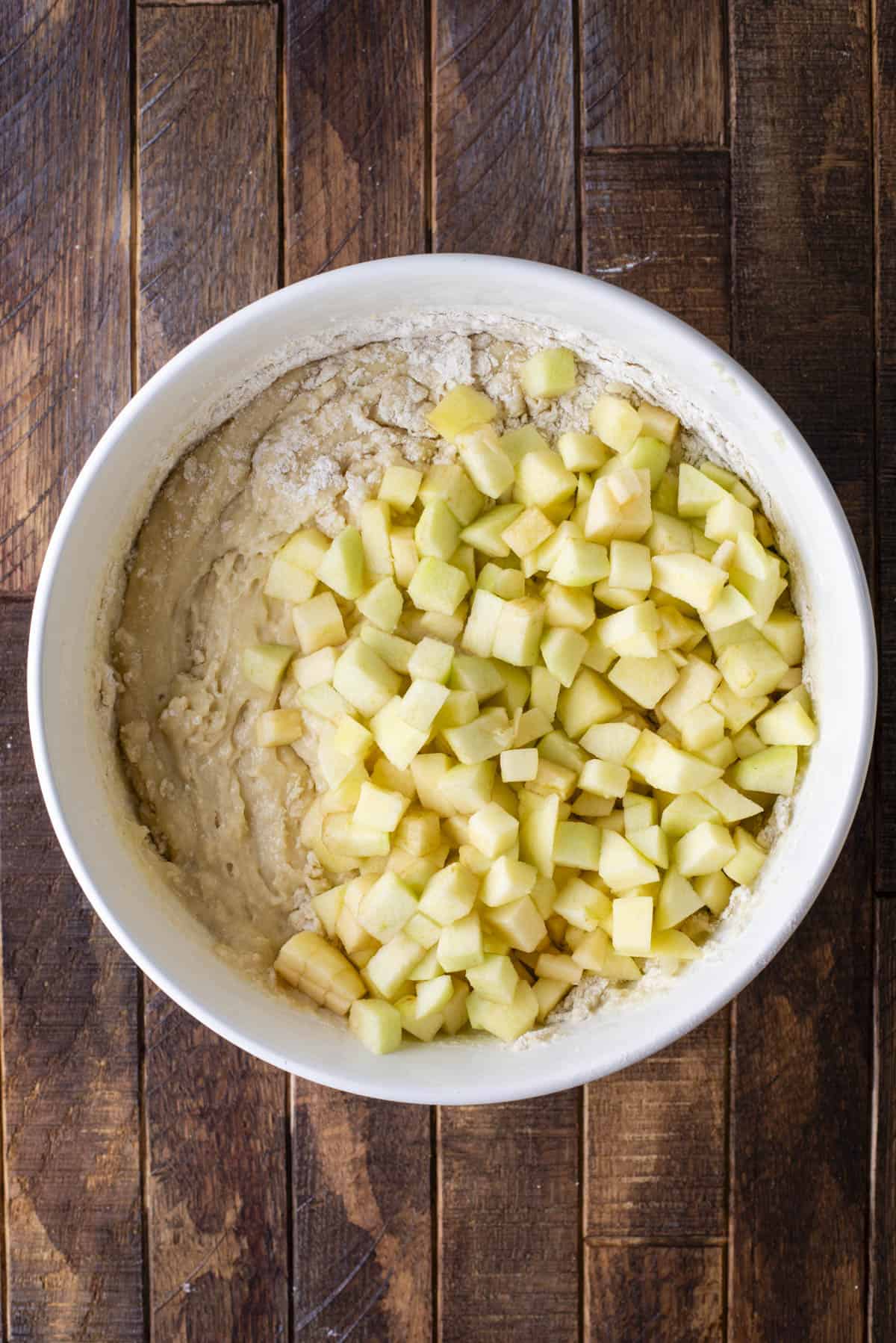 A white bowl containing muffin batter topped with a layer of chopped apple pieces, placed on a wooden surface.