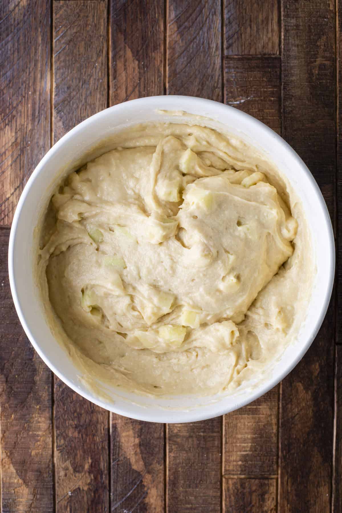 A white bowl filled with thick, creamy batter containing small chunks, sitting on a rustic wooden surface.