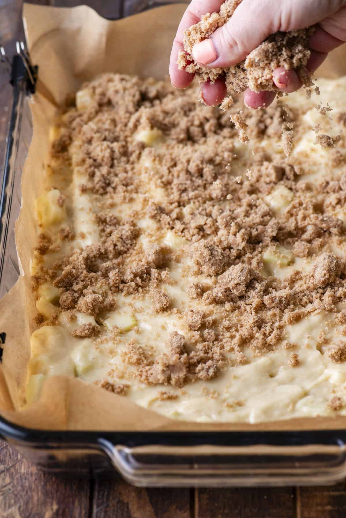 A hand sprinkles brown sugar topping over an unbaked cake batter in a parchment-lined glass baking dish on a wooden surface.