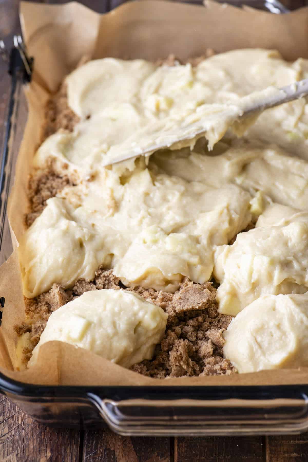 Close-up of a baking dish lined with parchment paper, filled with a crumbly brown sugar mixture being topped with spoonfuls of a creamy batter. A spatula is spreading the batter evenly over the crumb layer.