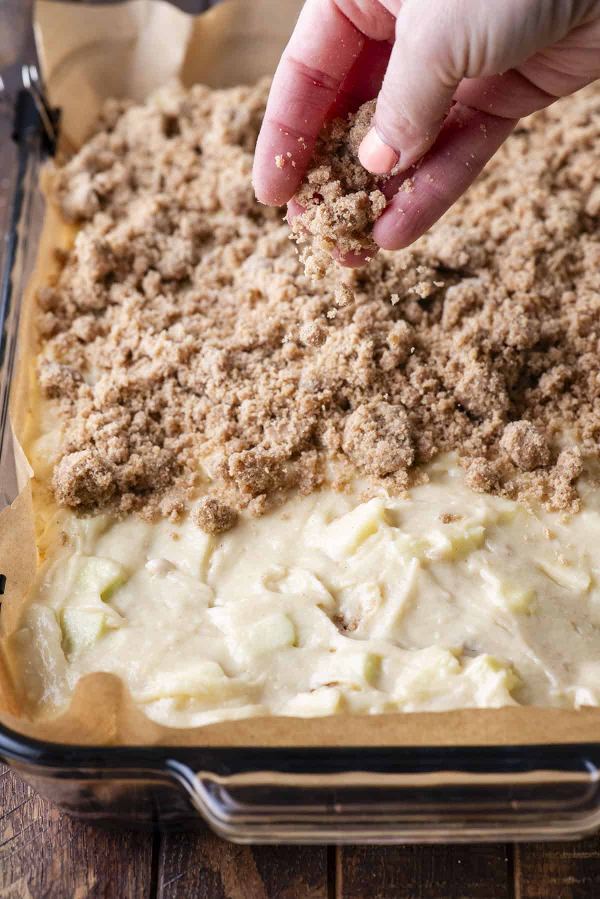 A hand sprinkles crumbly brown streusel topping over an unbaked apple cake batter in a parchment-lined baking dish.