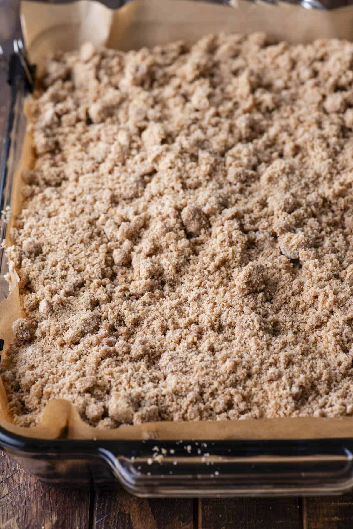 A glass baking dish lined with parchment paper, filled with an unbaked layer of crumbly streusel topping, ready to be baked.