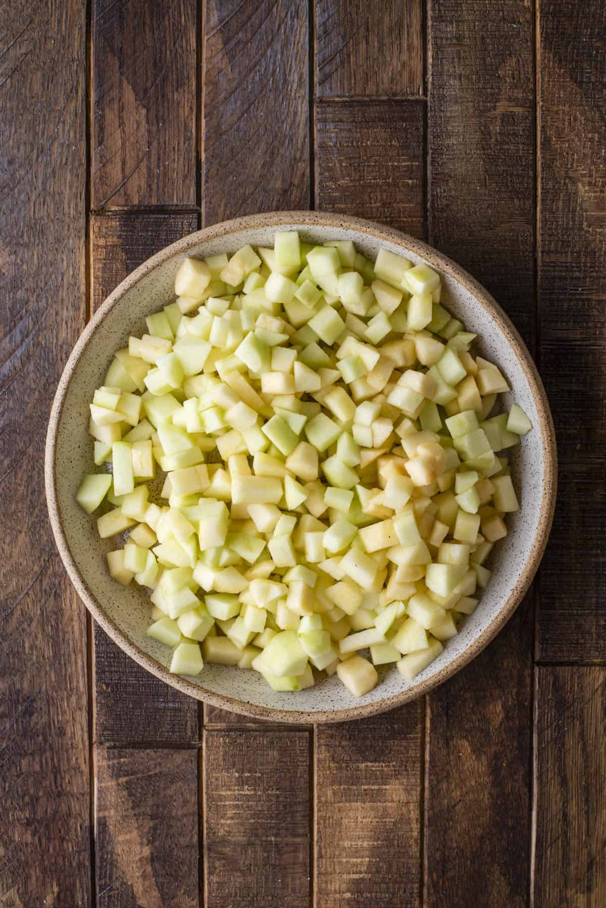 A ceramic bowl filled with small, evenly diced green apple pieces sits on a rustic wooden table.