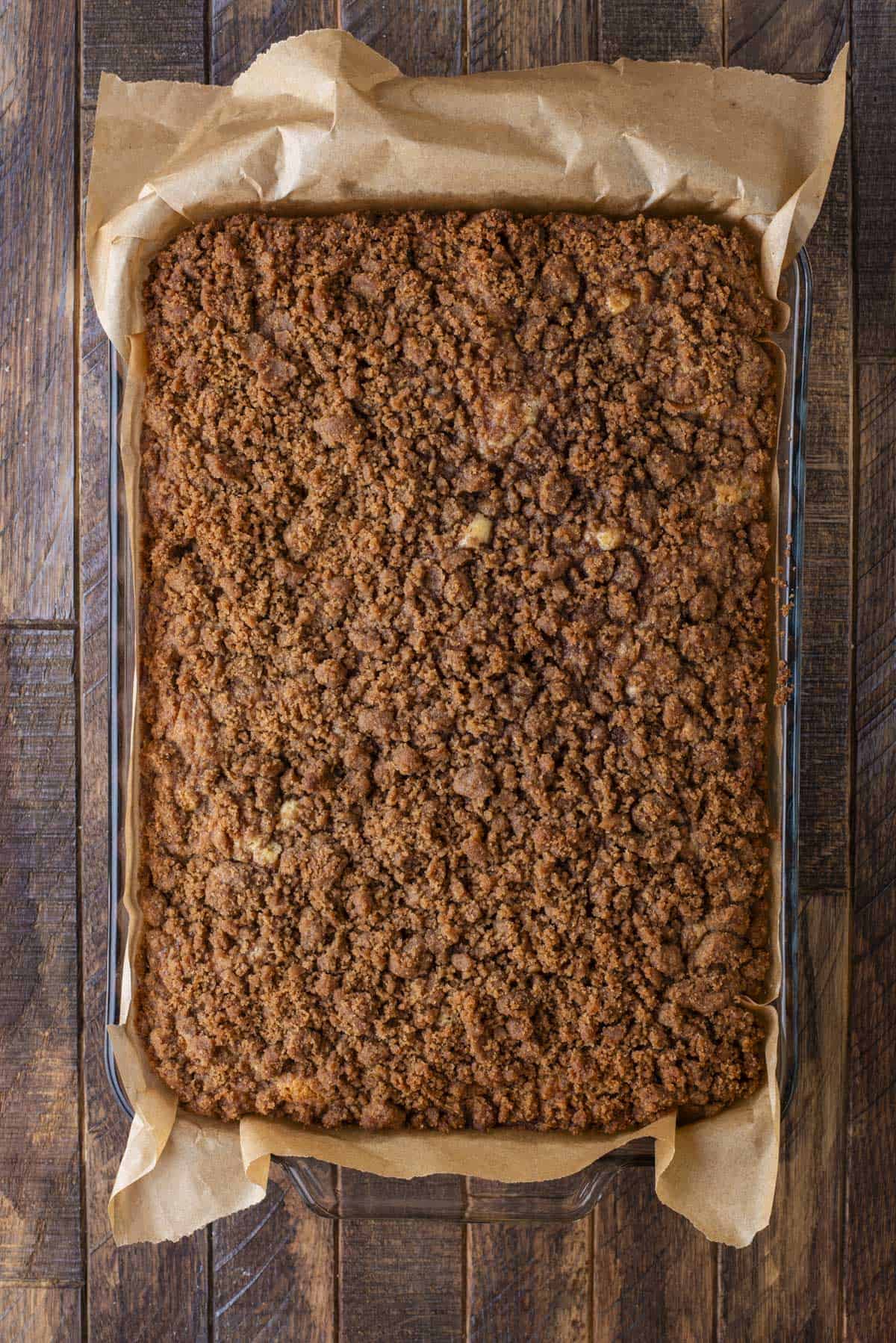 A rectangular glass baking dish lined with parchment paper holds a golden-brown crumb cake topped with a thick, crumbly cinnamon streusel. The dish sits on a rustic wooden table.