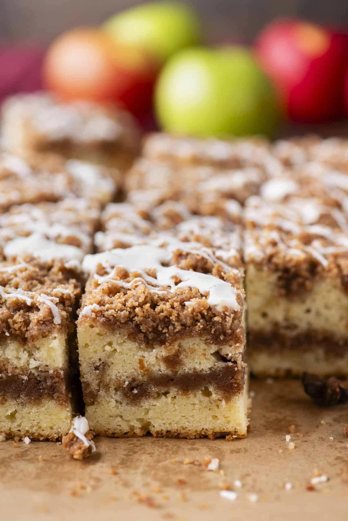 Close-up of sliced coffee cake with a crumbly cinnamon streusel topping and white icing drizzle, with blurred apples in the background.