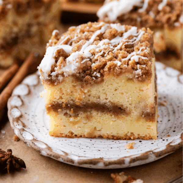 A slice of crumb-topped coffee cake with a cinnamon swirl filling and white icing drizzle, served on a speckled ceramic plate.
