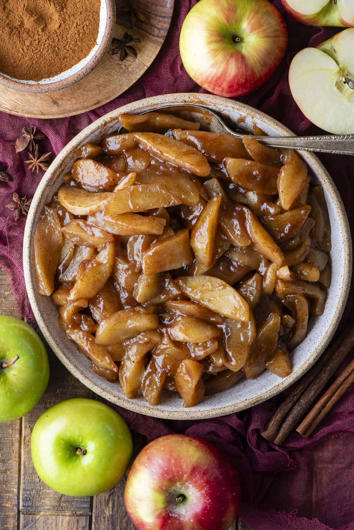 A bowl of cooked cinnamon apples with a spoon, surrounded by fresh red and green apples, cinnamon sticks, and a bowl of ground cinnamon on a rustic wooden surface.