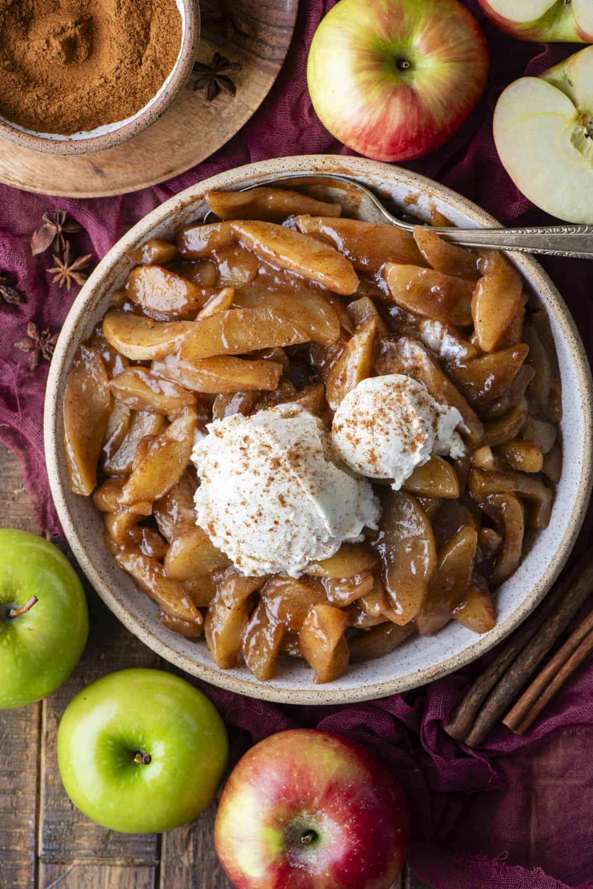 A bowl of baked cinnamon apples topped with two scoops of vanilla ice cream and sprinkled with cinnamon, surrounded by fresh apples, cinnamon sticks, and a bowl of ground cinnamon.