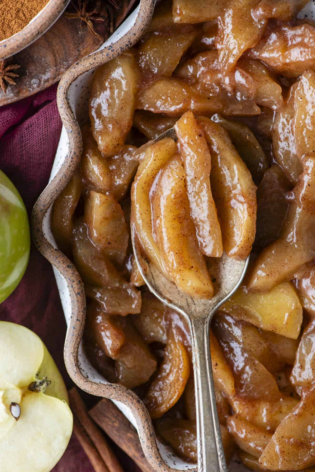 A close-up of baked cinnamon apples in a wavy-edged dish, with a spoon lifting several glazed apple slices. Green apples and cinnamon sticks are nearby.