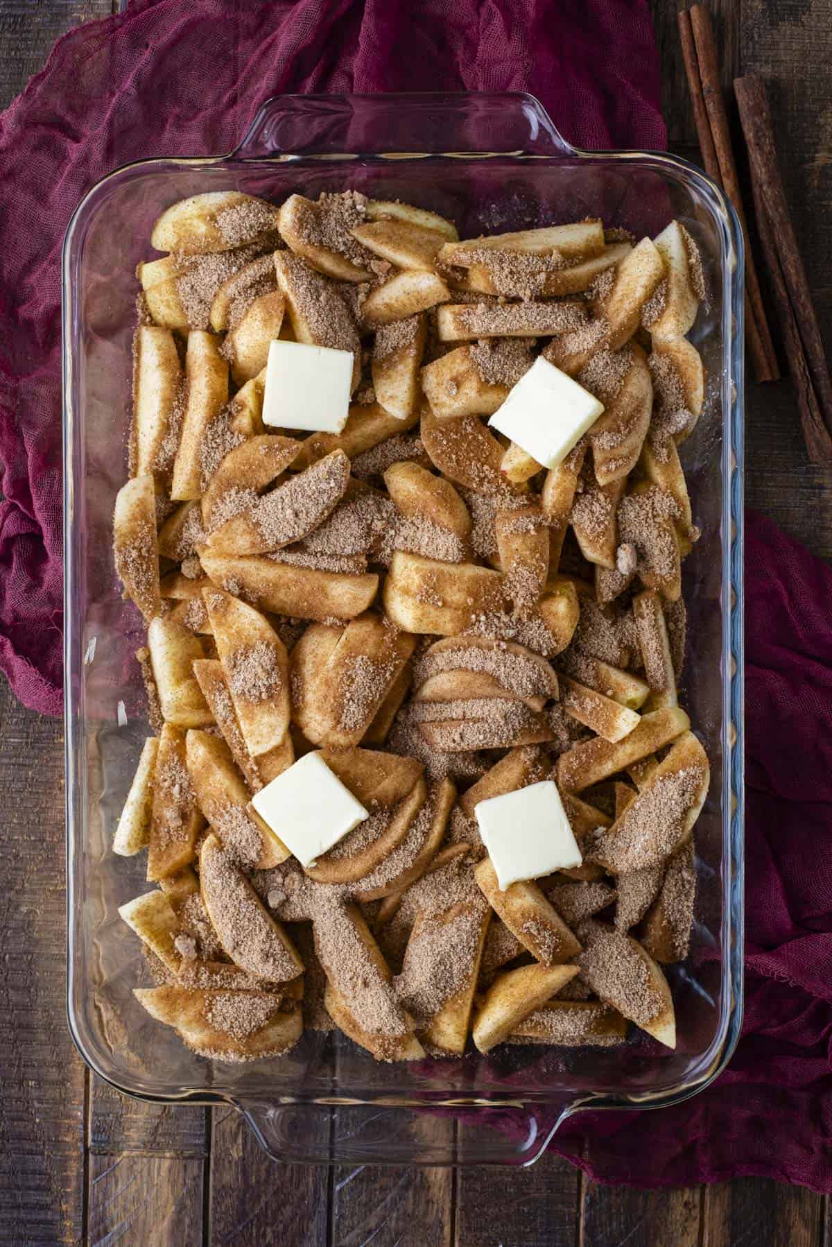 A glass baking dish filled with sliced apples topped with a layer of cinnamon sugar and four cubes of butter, set on a rustic wooden surface with a deep red cloth and cinnamon sticks beside it.