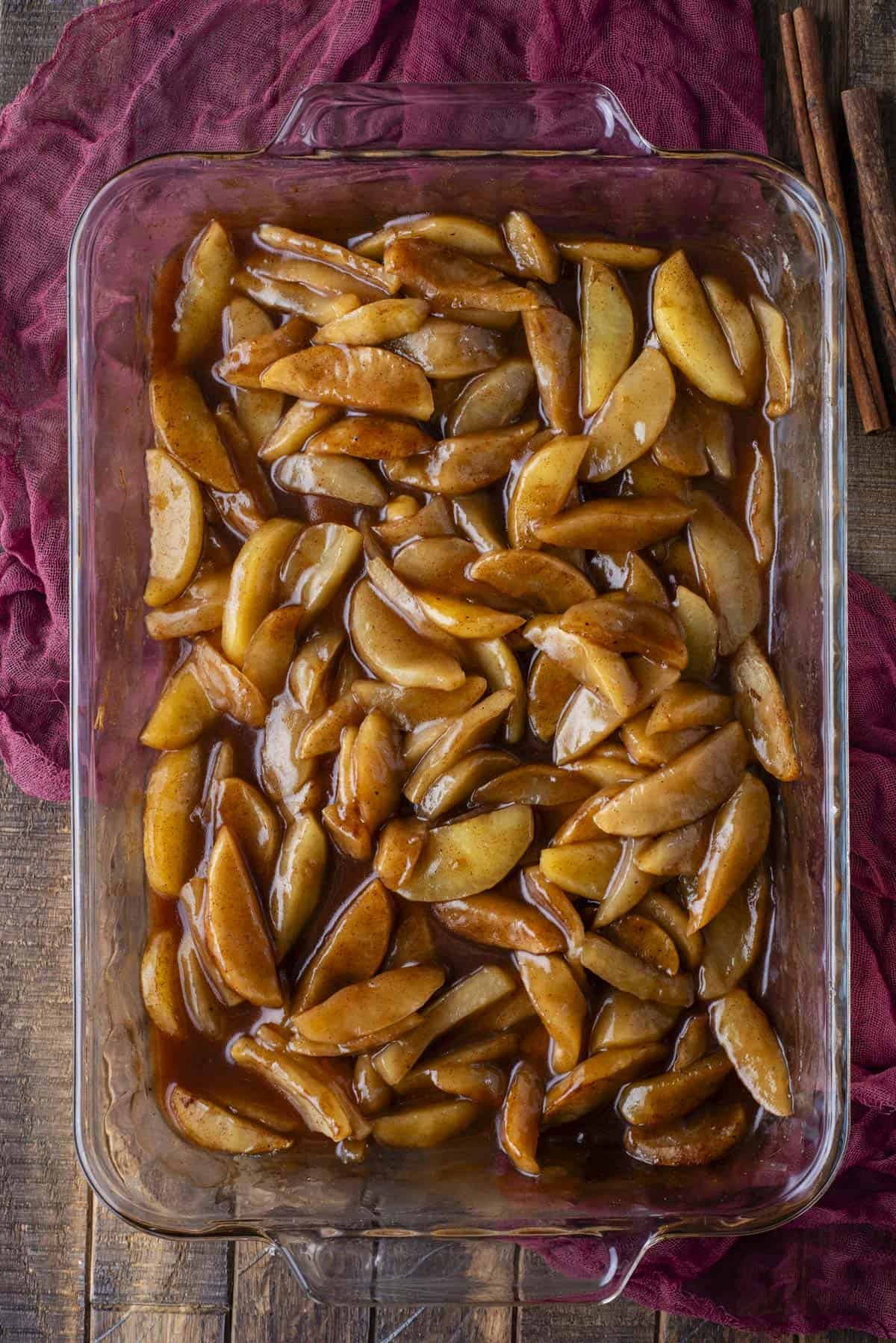 A glass baking dish filled with sliced apples coated in a glossy cinnamon sauce, sitting on a wooden surface with a dark red cloth and cinnamon sticks beside it.