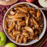 A bowl filled with baked apples coated in a glossy brown cinnamon sauce sits on a rustic surface, surrounded by green apples, cinnamon sticks, and star anise. A silver spoon rests in the bowl.