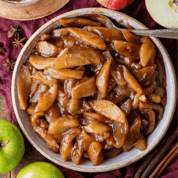 A bowl filled with baked apples coated in a glossy brown cinnamon sauce sits on a rustic surface, surrounded by green apples, cinnamon sticks, and star anise. A silver spoon rests in the bowl.