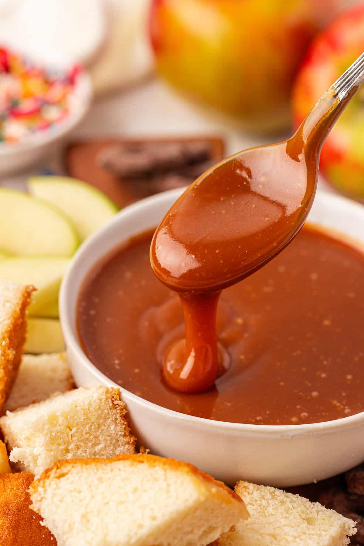 A spoon drizzles caramel sauce into a white bowl, surrounded by cubes of cake, green apple slices, and colorful sprinkles in the background.