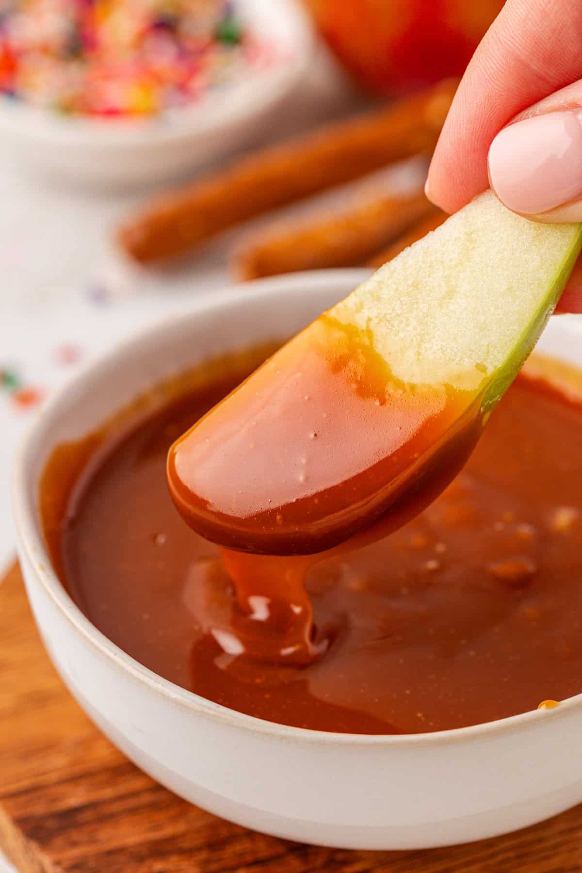 A hand dips a fresh apple slice into a bowl of smooth caramel sauce, with cinnamon sticks and colorful sprinkles blurred in the background.