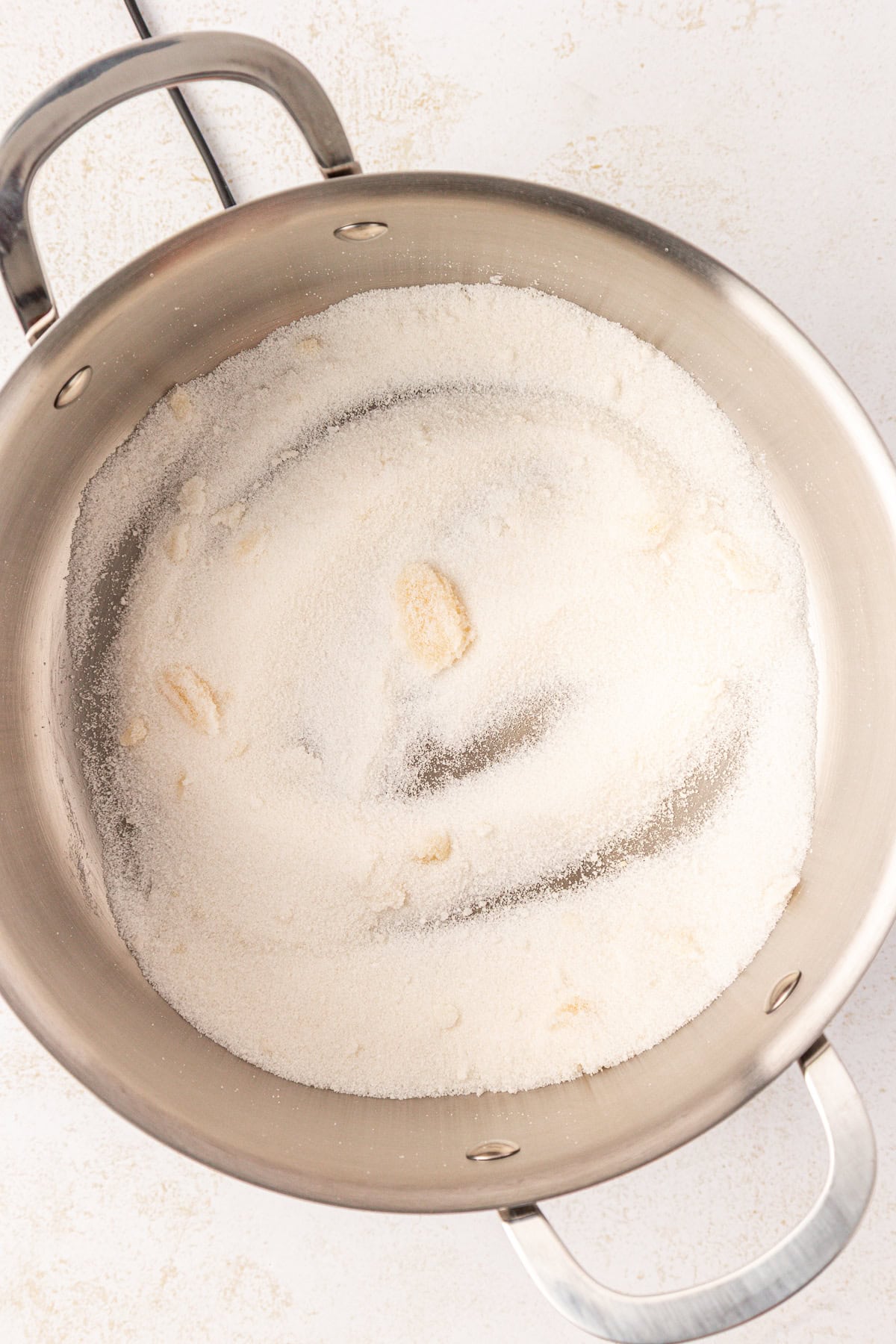 A stainless steel pot containing a layer of granulated sugar with a few clumps, sitting on a light-colored surface.