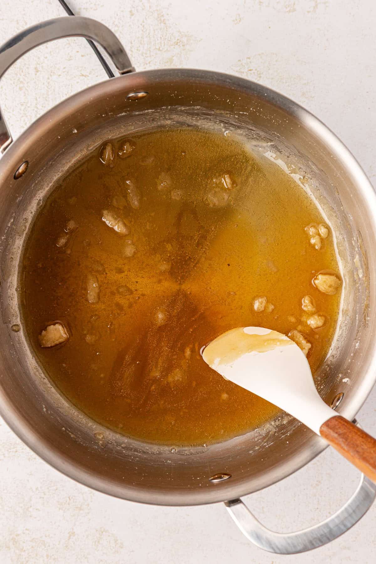 A stainless steel pot containing golden brown melted sugar with a few lumps, being stirred by a white spoon with a wooden handle, sits on a light-colored surface.