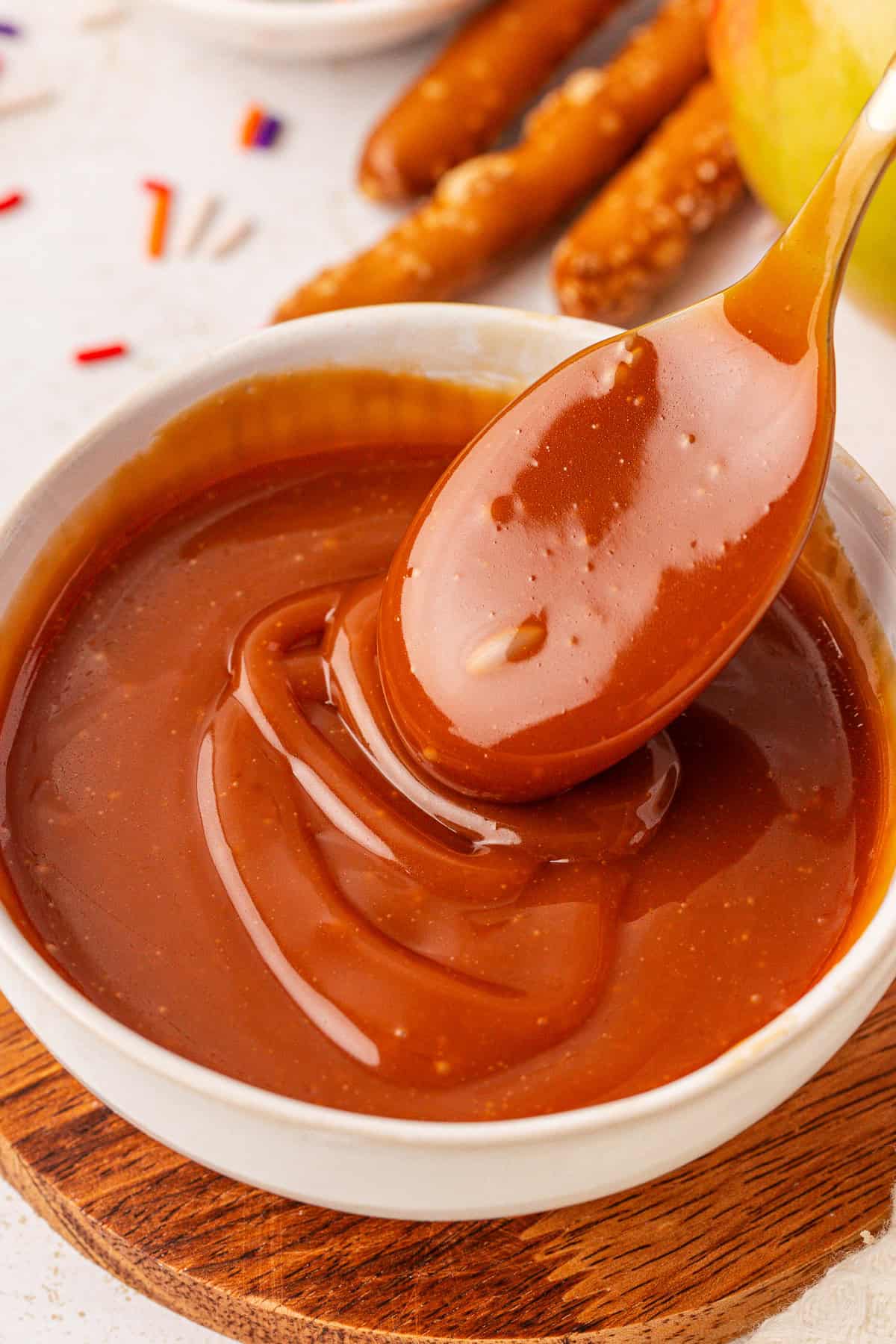 A spoon lifts creamy caramel sauce from a white bowl, with pretzel sticks and apple slices blurred in the background.