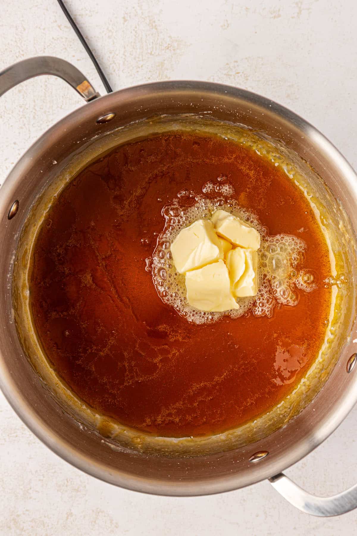 A stainless steel pot containing melted brown sugar with several chunks of butter starting to melt in the center. The mixture has a glossy, caramel-like appearance.