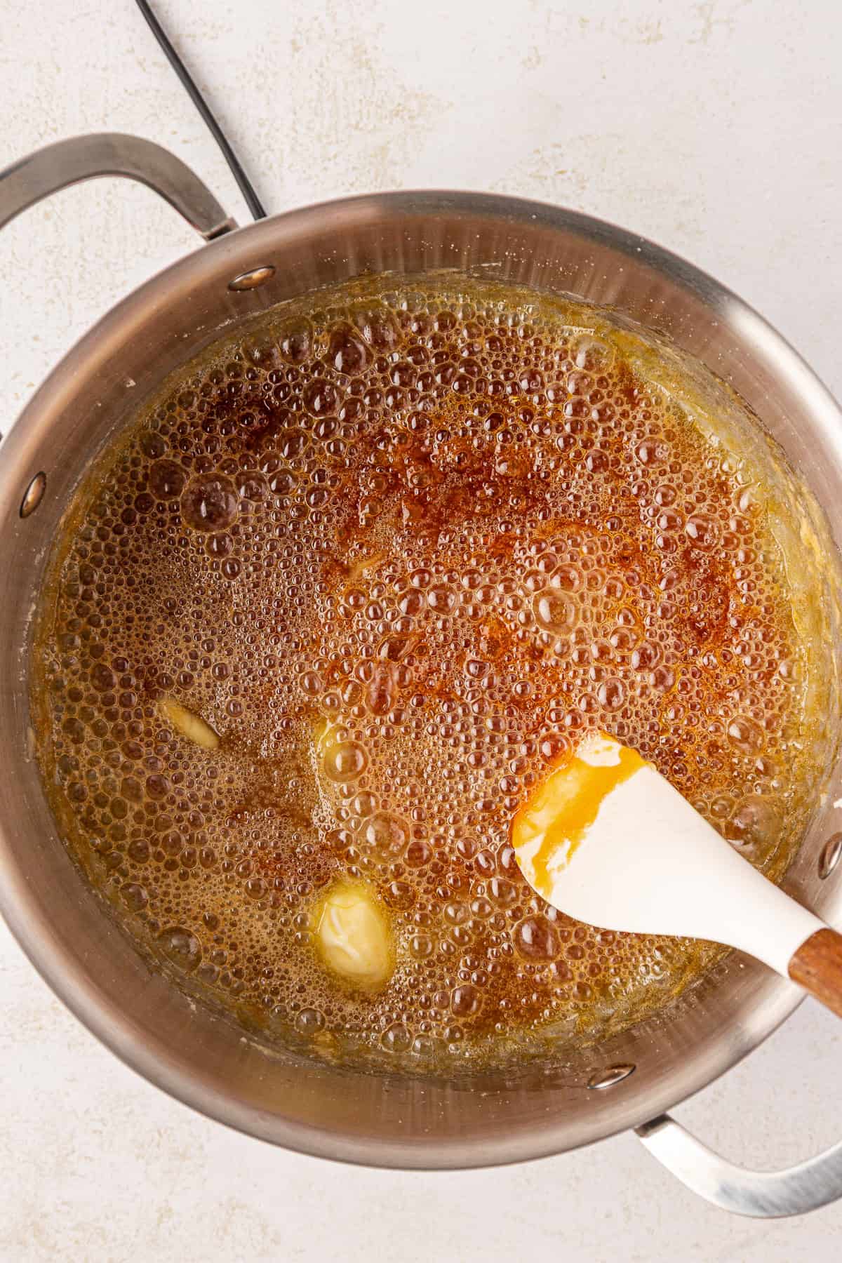 A saucepan filled with bubbling brown caramel and melting butter, being stirred with a white and wooden spatula on a light countertop.