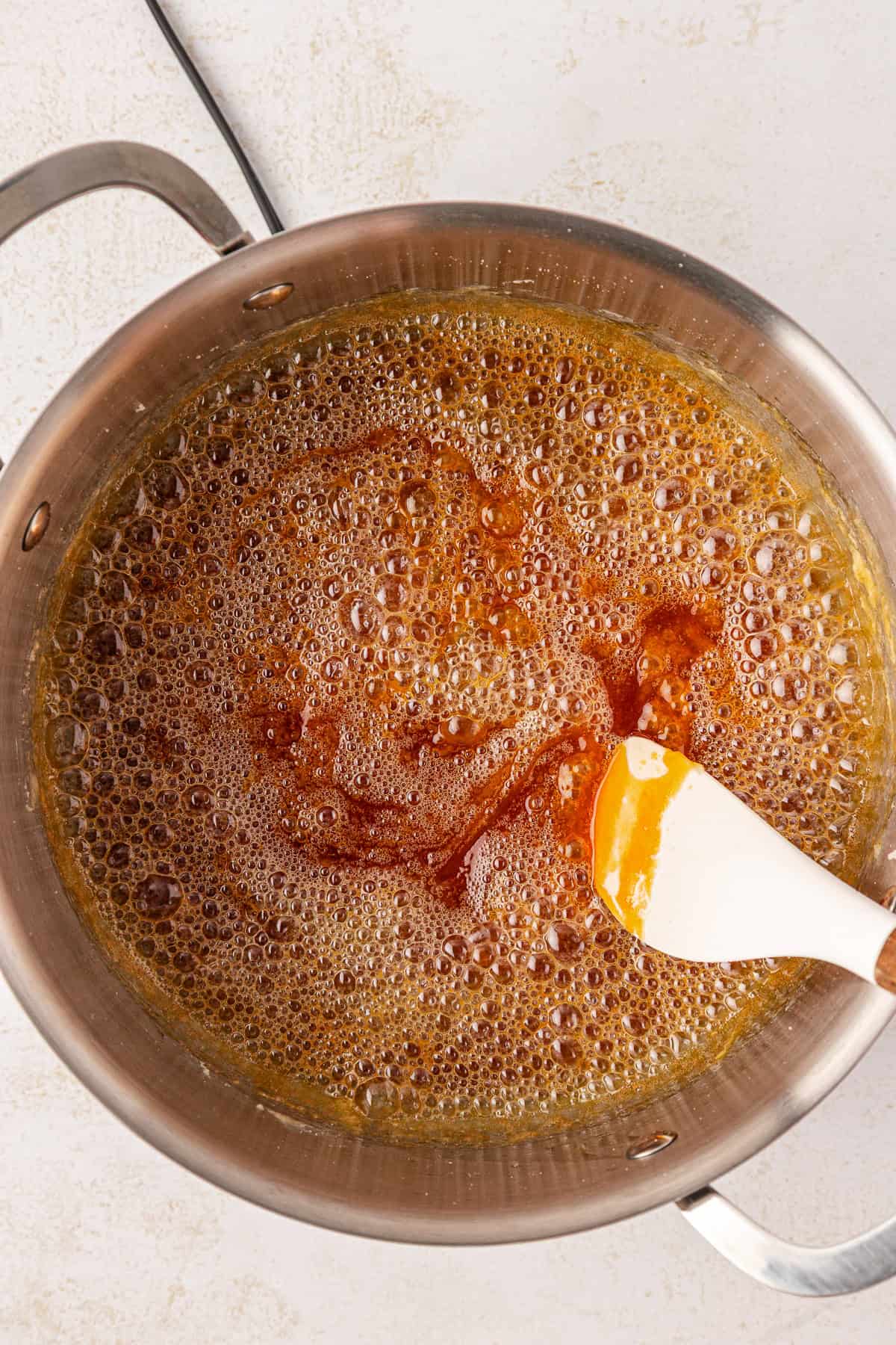 A saucepan filled with bubbling amber-colored caramel is being stirred with a white spatula. The pot handles are visible on both sides, and the scene is set on a light surface.
