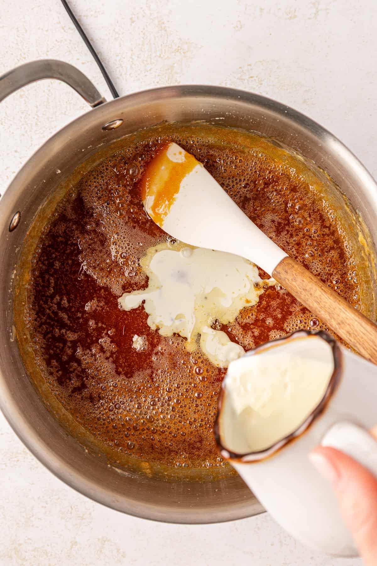 A hand pours cream from a small cup into a saucepan of bubbling caramel, with a white spatula resting inside the pan.