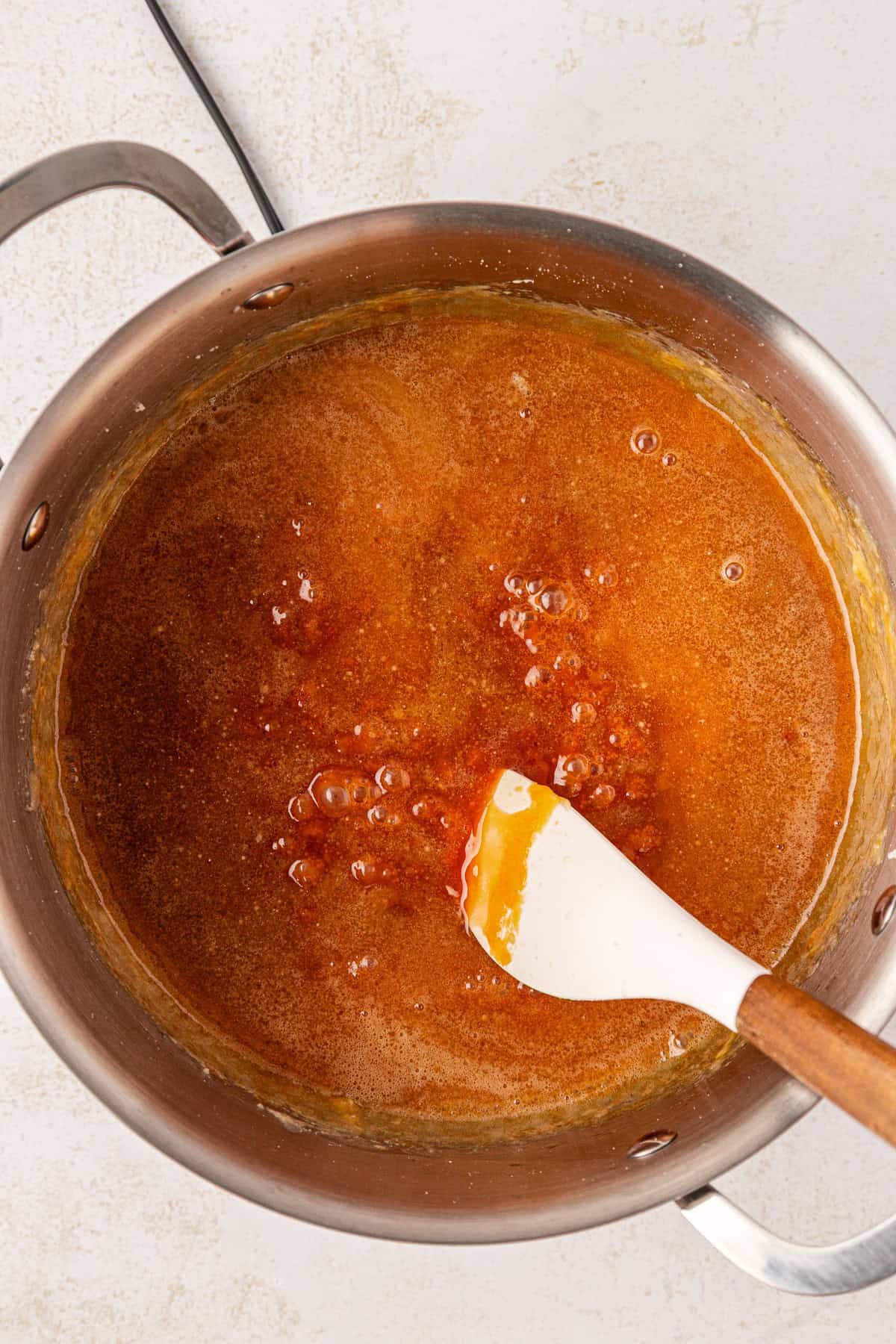 A saucepan filled with bubbling caramel sauce is being stirred with a white and wooden spatula, shown from above on a light-colored countertop.