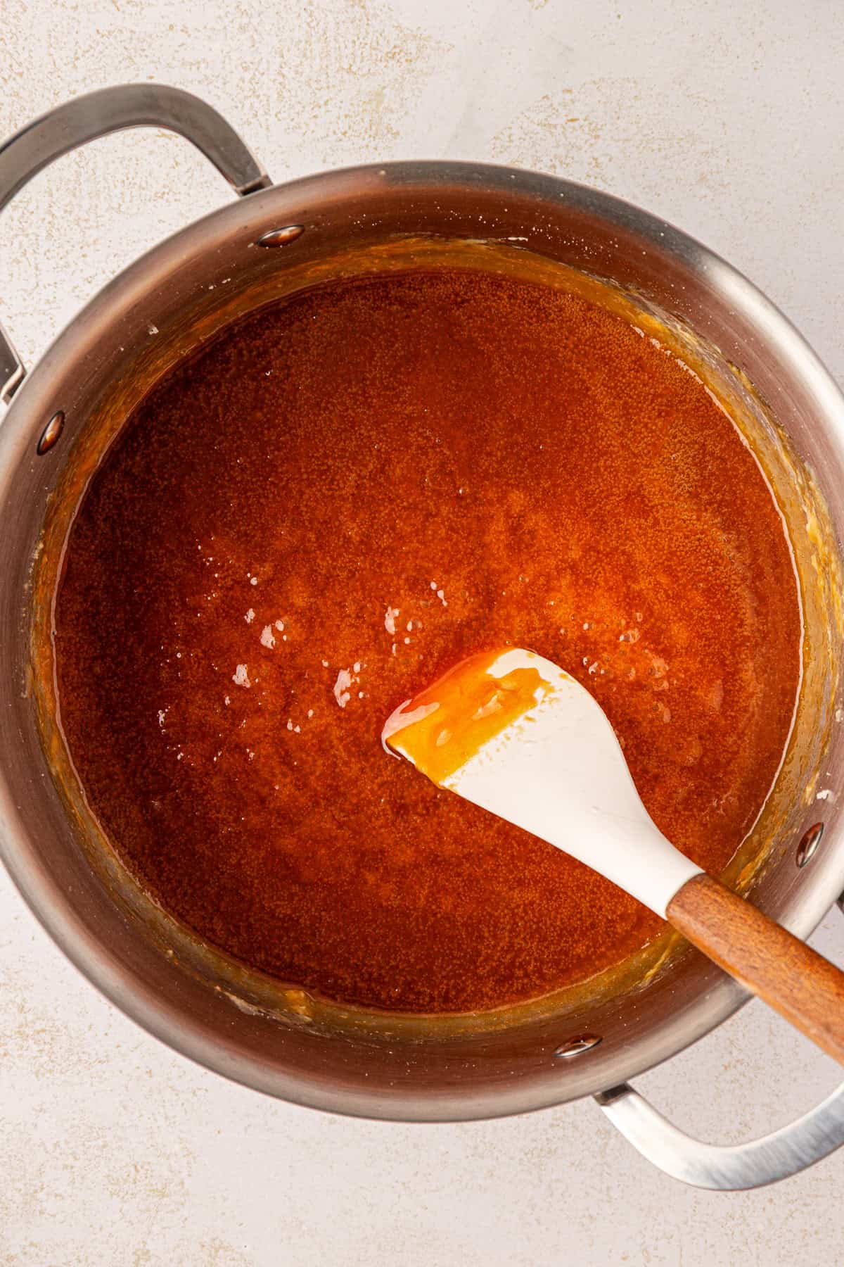 Overhead view of a saucepan filled with golden-brown caramel sauce. A white silicone spatula with a wooden handle rests inside, coated with the sauce. The pan sits on a light-colored countertop.