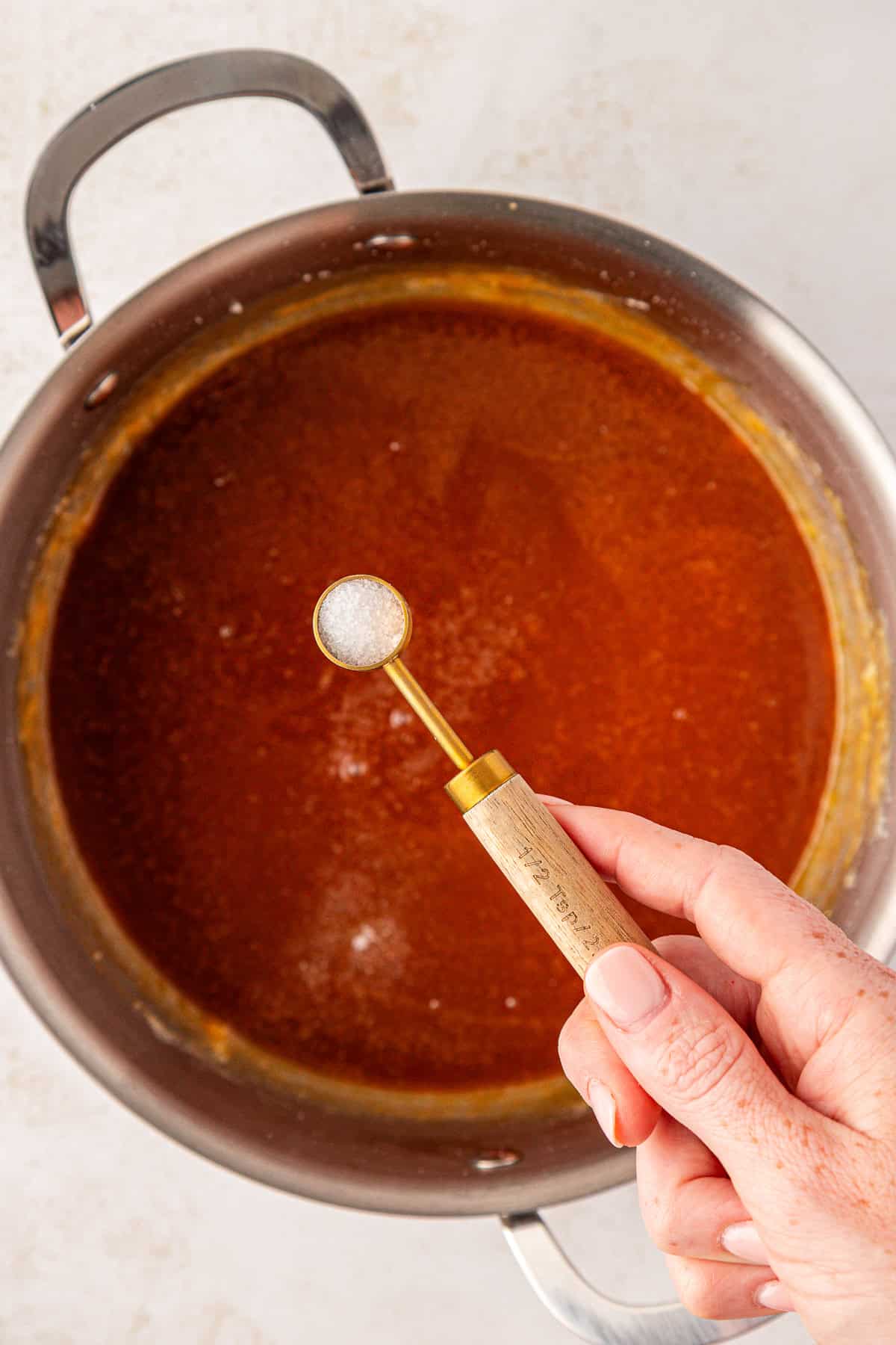 A hand holding a small measuring spoon of salt over a pot filled with a red-orange sauce or soup.