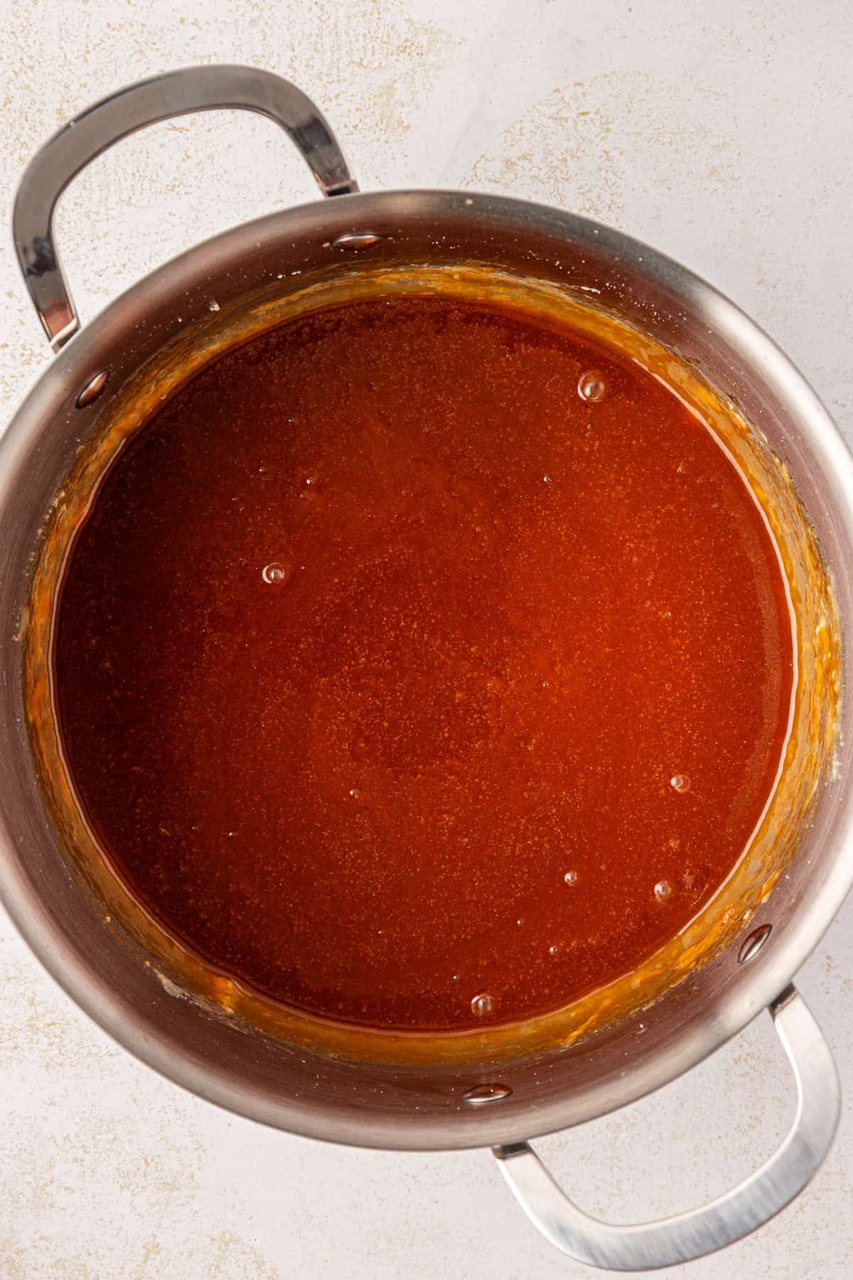 A large stainless steel pot filled with smooth, rich brown sauce sits on a white surface, with sauce residue visible along the inner sides of the pot.