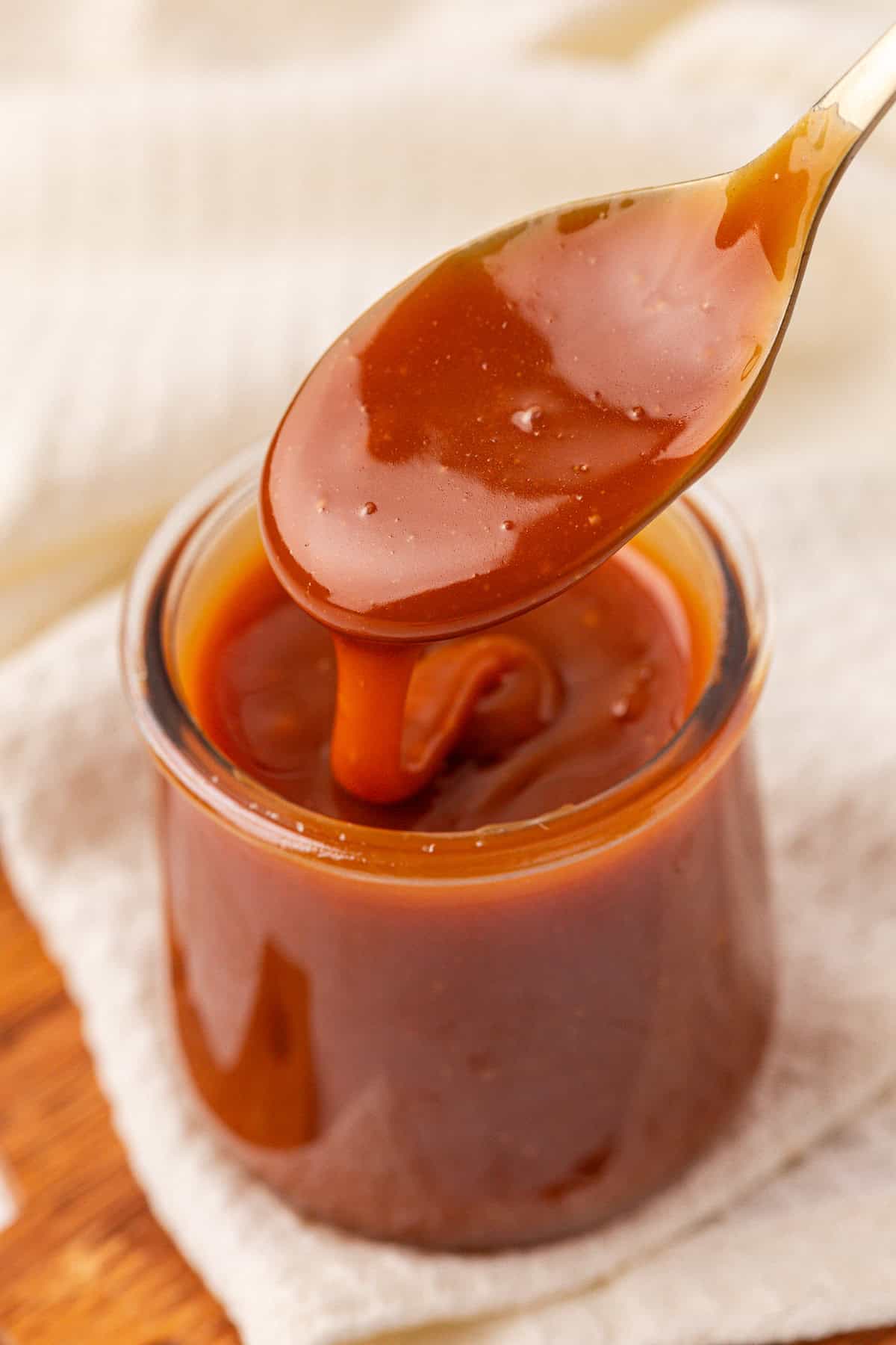 A close-up of a spoon dipped in a glass jar filled with thick, glossy caramel sauce, with the sauce dripping off the spoon. The jar sits on a light-colored cloth.