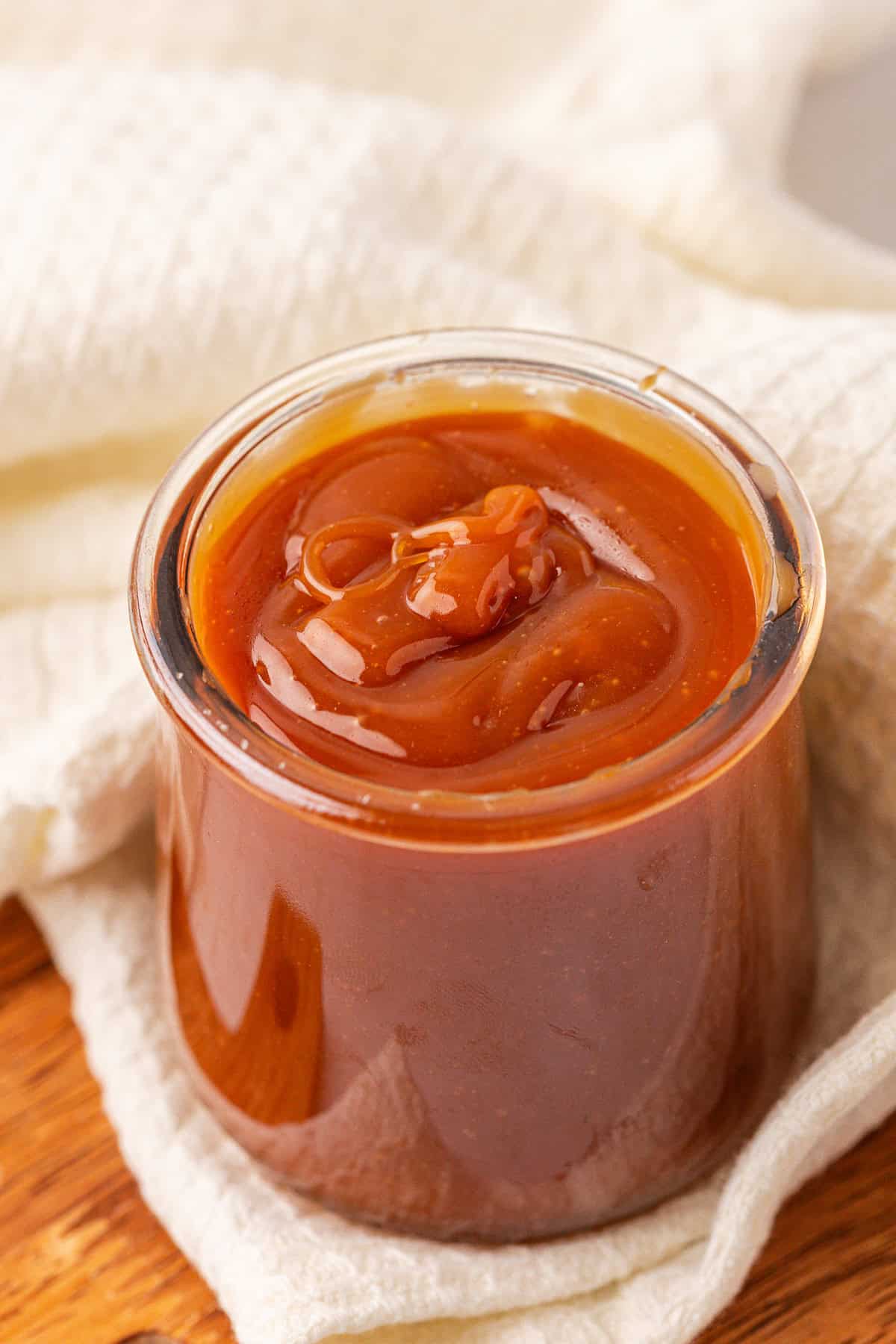 A small glass jar filled with smooth, rich caramel sauce sits on a white cloth atop a wooden surface. The caramel has a glossy texture and is swirled on top.