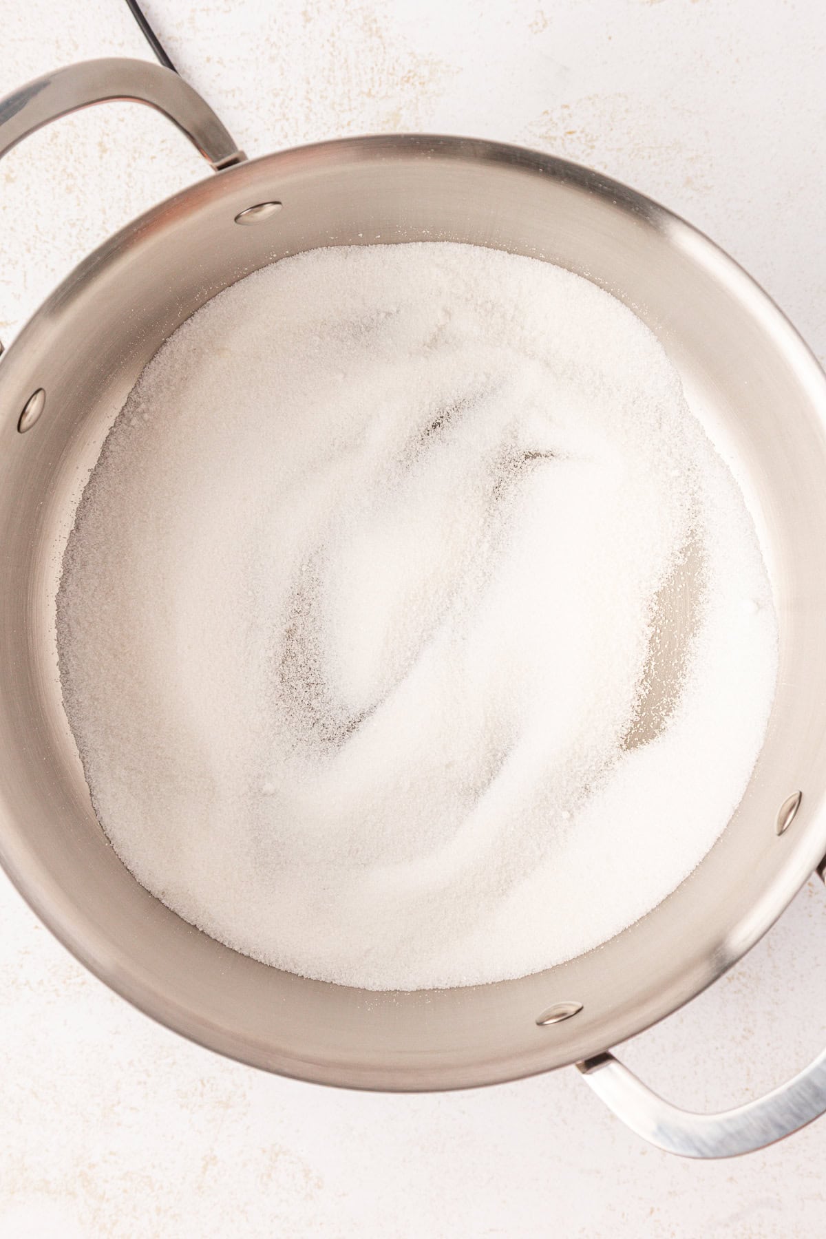 A close-up of a stainless steel pot filled with a layer of white granulated sugar, placed on a light-colored surface.